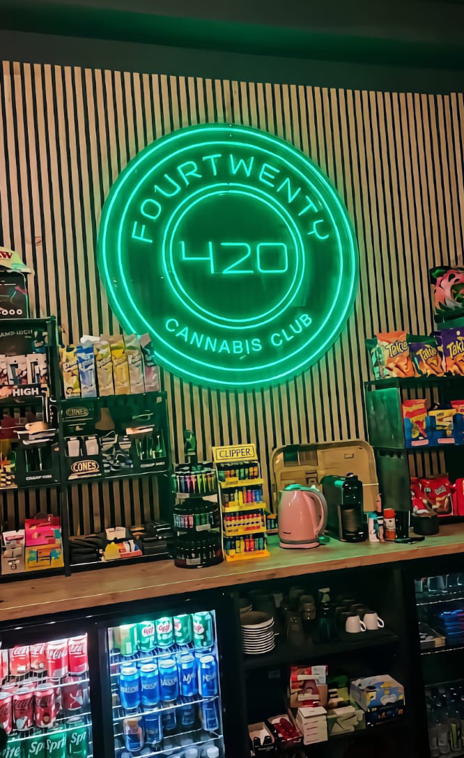 A bright green neon sign with the logo at Four Twenty cannabis club in Valencia, displayed above a bar counter filled with snacks, drinks, and Clipper lighters.