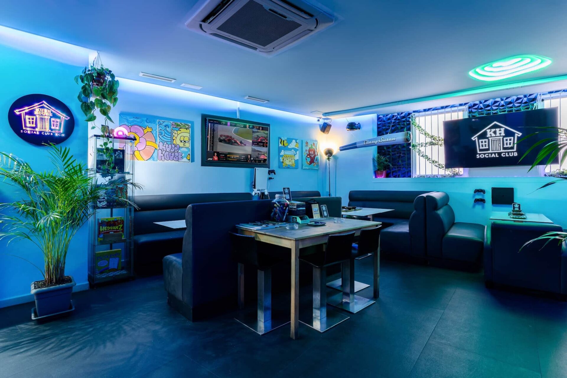 Interior of Kush House Social Club in Barcelona, with blue ambient lighting, booth seating, gaming screens, colorful wall art, and potted plants