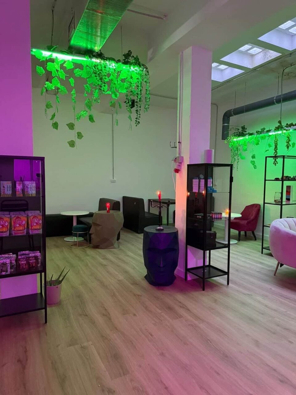 Interior of Mística Social Club in Madrid, showing modern seating, display shelves, green neon lights, and hanging plants.
