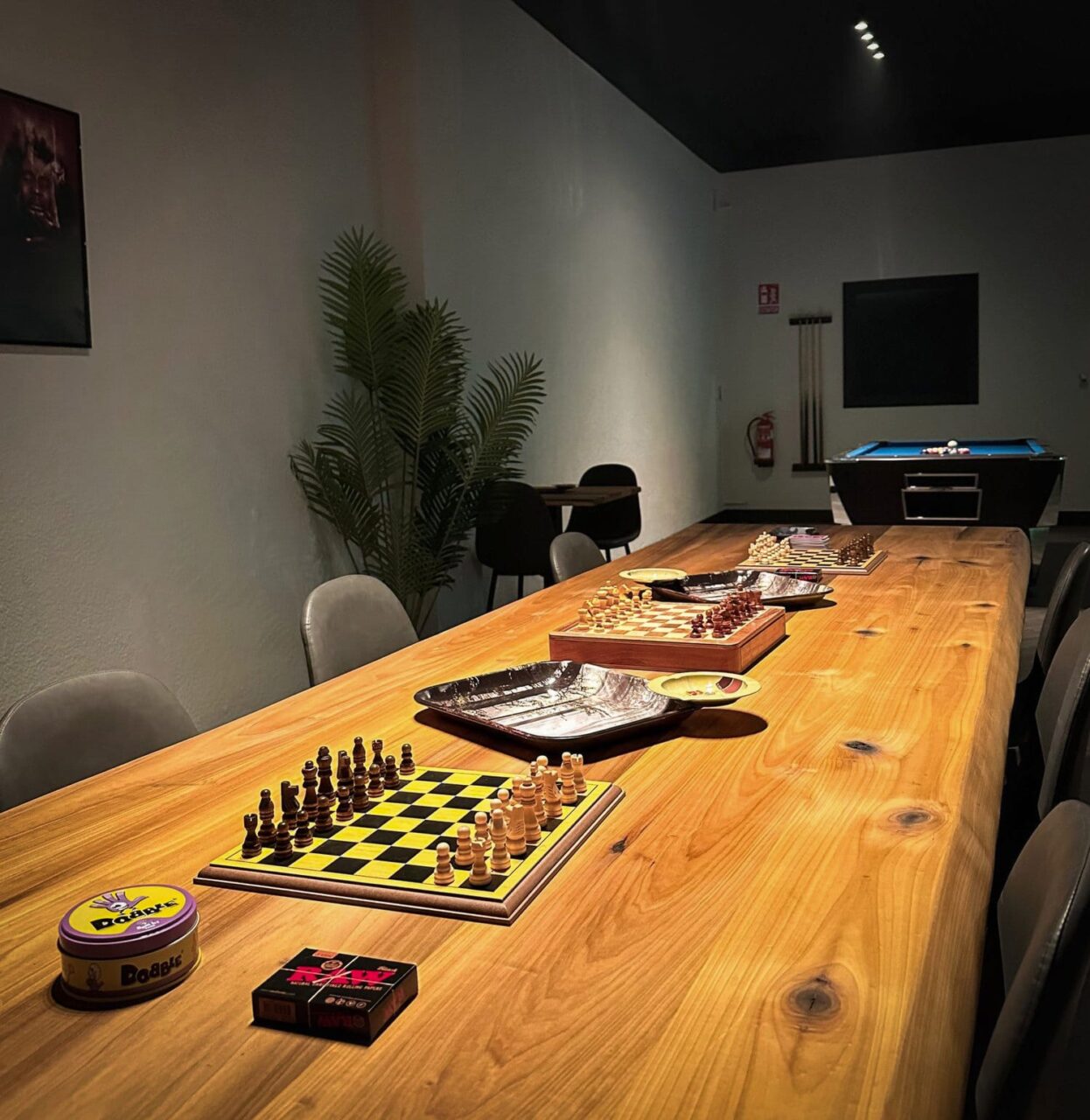 A long wooden table set with chess boards, board games, and smoking accessories at Four Twenty cannabis club in Valencia, with a pool table visible in the background.