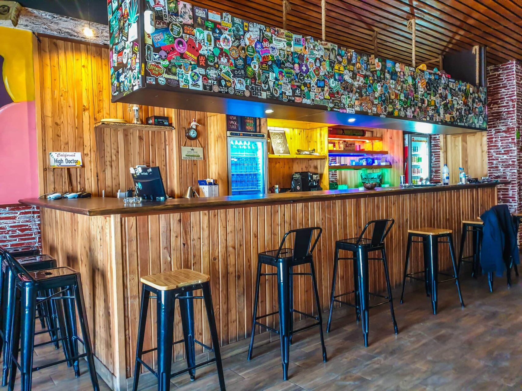 Interior of The High Doctor CSC bar in Alicante with wooden counter, colorful beer can decor and metal stools.