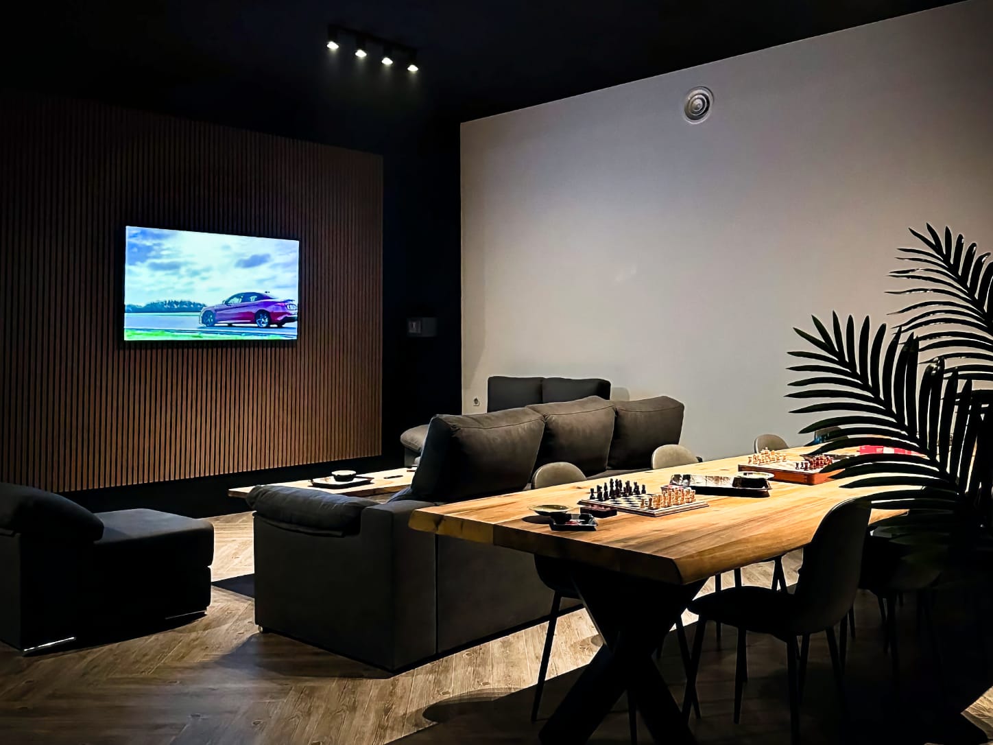 The cozy lounge area at Four Twenty cannabis club in Valencia, featuring dark sofas facing a large TV screen on a wooden slat wall, and chess sets on a long wooden table.