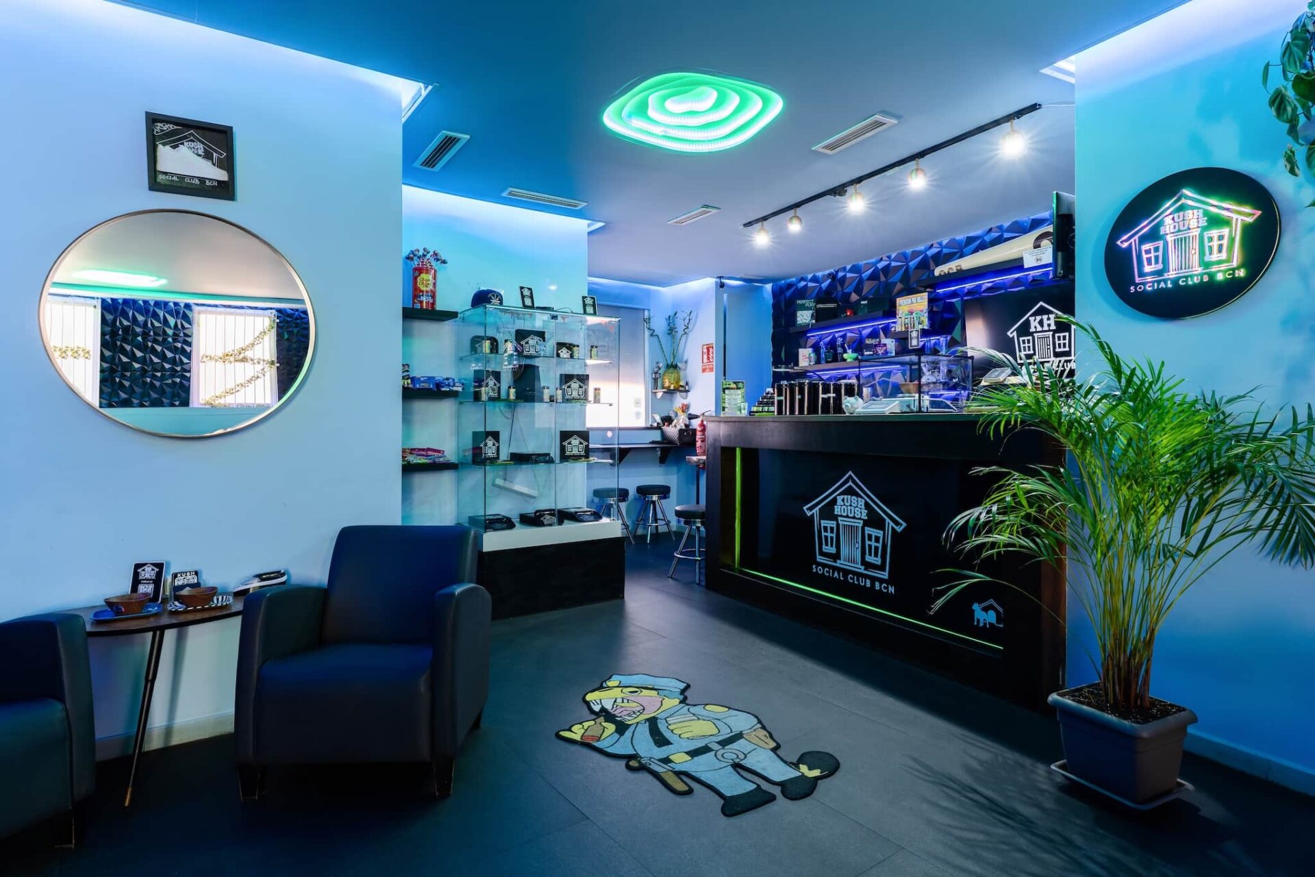 Reception area of Kush House Social Club in Barcelona, with a modern counter, blue ambient lighting, product display shelves, armchairs, and a cartoon rug on the floor
