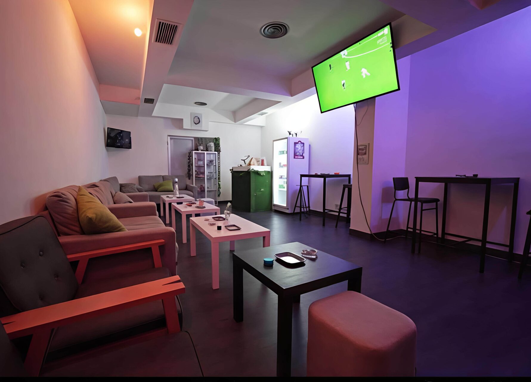 Lounge area at Valhalla cannabis club in Madrid with seating, high tables, a cooler, and a TV showing a football match.