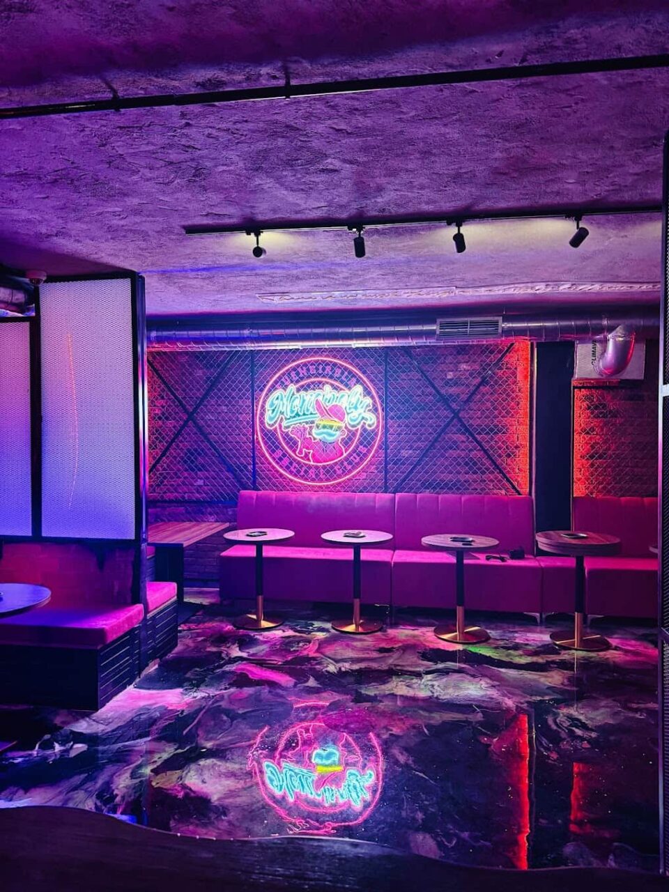 Lounge area of Monopoly Social Club in Malaga, with purple ambient lighting, brick wall backdrop, pink upholstered sofas, and reflective floor.